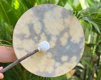 May include: A hand-crafted drum with a natural leather surface and a wooden beater. The drum has a mottled gray and beige pattern. The drum is being held by a person's hand.