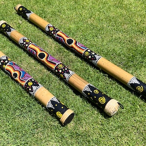 May include: Three wooden didgeridoos with colorful, intricate designs painted on them. The didgeridoos are lying on a green grassy surface.