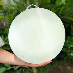 Op de afbeelding: Een cirkelvormige, gebroken witte trommel met een glad, strak oppervlak en een kleine witte lus aan de bovenrand om op te hangen. De trommel heeft een consistente, bleke, matte afwerking over de gehele voorzijde.