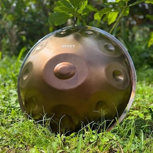May include: A bronze-colored steel handpan instrument with the word "LIDAH" printed on the surface. The instrument has several circular indentations and is set against a backdrop of green grass and foliage.
