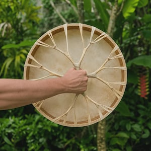 Pode incluir: Um tambor de moldura circular com uma pele opaca de cor castanho-claro feita de pele esticada. O tambor apresenta um aro de madeira e um sistema complexo de cordões finos e claros que irradiam do centro para a moldura exterior.