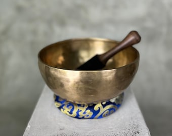 May include: A metallic gold-colored singing bowl with a smooth, rounded exterior and a dark wooden mallet resting inside. The bowl sits on a circular blue fabric cushion featuring a repeating gold-colored pattern of swirling designs and symbols.