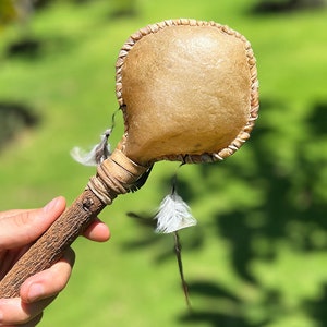 Pode incluir: Um instrumento de percussão artesanal, castanho claro, com uma cabeça arredondada e costurada e um cabo de madeira texturizado. Penas brancas são presas perto da cabeça. O instrumento é segurado contra um fundo verde desfocado.