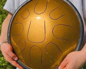 Könnte beinhalten: Ein goldfarbenes Handpan mit schwarzem Rand. Das Handpan hat eine runde Form und ist mit einem Muster aus Vertiefungen verziert.