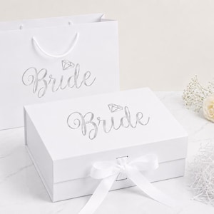 May include: A white gift bag and a rectangular white gift box, both featuring the word Bride in silver script lettering with a small diamond icon above the text. The gift box includes a white ribbon closure tied in a bow at the front center.