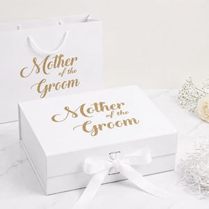May include: A white gift bag and a rectangular white gift box, both featuring the gold text Mother of the Groom. The box has a hinged lid secured with a white ribbon tied in a bow. The set is accompanied by two white roses and white paper shred.