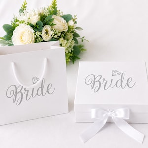 May include: A white gift bag and a square white gift box, both featuring the word Bride in silver script with a small diamond icon above it. The box is adorned with a white satin ribbon bow, and a bouquet of white roses and greenery sits behind.