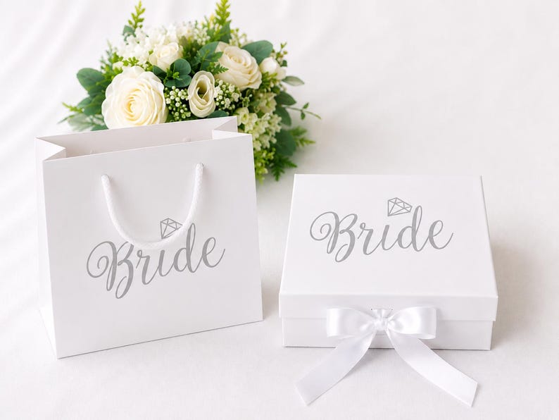 May include: A white gift bag and a square white gift box, both featuring the word Bride in silver script with a small diamond icon above it. The box is adorned with a white satin ribbon bow, and a bouquet of white roses and greenery sits behind.