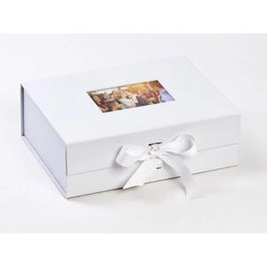 May include: A rectangular white gift box featuring a hinged lid with a central rectangular window displaying a colorful photograph of a group of people. The box is secured with a white ribbon tied into a bow on the front panel of the container.