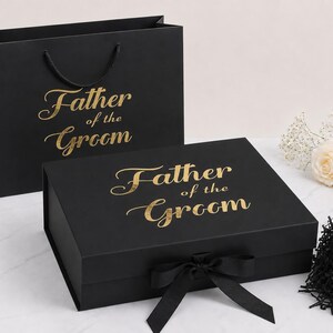May include: A black gift bag and a rectangular black gift box, both featuring the gold foil text Father of the Groom. The box has a black ribbon closure at the front. The set is accompanied by a small pile of black shredded paper filler.