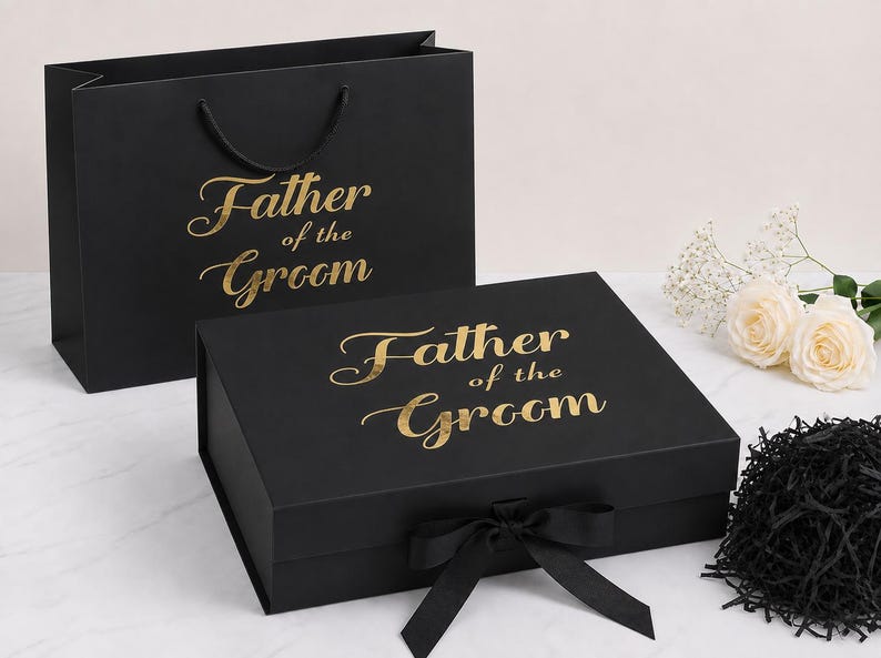 May include: A black gift bag and a rectangular black gift box, both featuring the gold foil text Father of the Groom. The box has a black ribbon closure at the front. The set is accompanied by a small pile of black shredded paper filler.