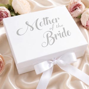 Pode incluir: Uma caixa de presente retangular branca com o texto prateado "Mother of the Bride" impresso na tampa. A caixa é fechada com uma fita de cetim branca atada num laço na frente, decorada com várias flores de peónia cor-de-rosa suave.