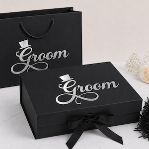 May include: A black gift bag and a matching black rectangular gift box, both featuring the word Groom in silver script with a small top hat icon above the letter G. The box includes a black ribbon closure and black shredded paper filler.