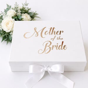 Może przedstawiać: Prostokątne białe pudełko prezentowe z napisem Mother of the Bride wykonanym metaliczną złotą czcionką na wieczku. Pudełko jest zamknięte białą satynową wstążką zawiązaną w kokardę na środku i ozdobione małą kompozycją kwiatową.