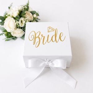 May include: A square white gift box featuring the word Bride in gold cursive script with a small gold diamond icon above it. The box is closed with a white satin ribbon tied in a bow at the front, accompanied by a bouquet of white roses and greenery.