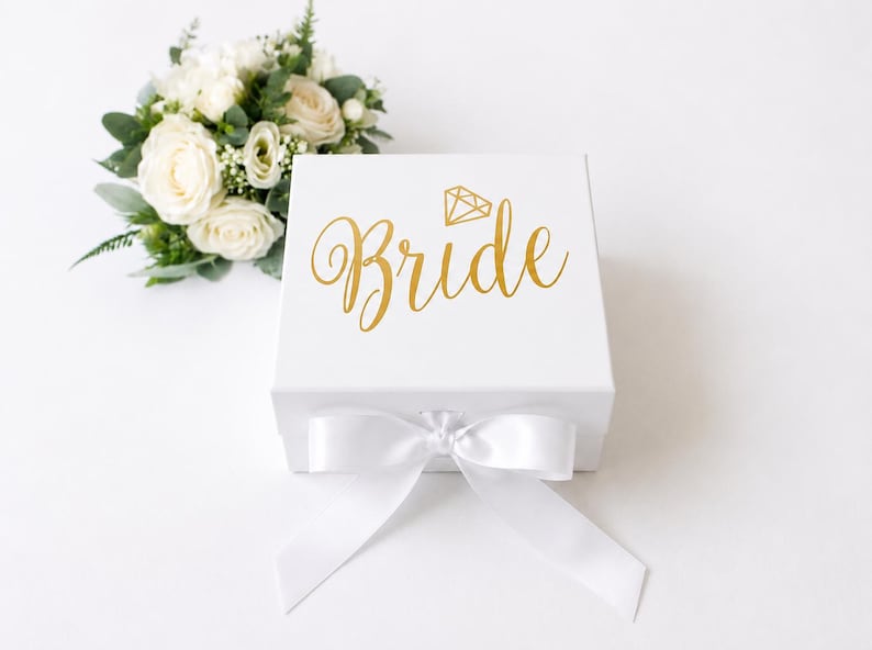 May include: A square white gift box featuring the gold text Bride and a small gold diamond icon on the lid. The box is closed with a white satin ribbon tied in a bow at the front, accompanied by a small bouquet of white roses and green foliage.