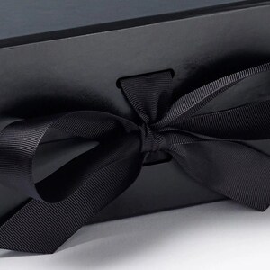 May include: A matte black rectangular gift box featuring a smooth surface and a centered horizontal slot. A wide, black grosgrain ribbon is threaded through the slot and tied into a decorative bow on the front exterior of the box.