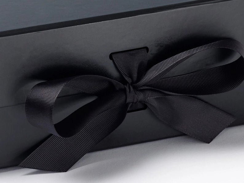 May include: A matte black rectangular gift box featuring a smooth surface and a centered horizontal slot. A wide, black grosgrain ribbon is threaded through the slot and tied into a decorative bow on the front exterior of the box.