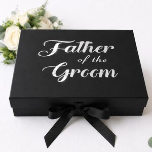 May include: A rectangular black gift box featuring the silver text Father of the Groom printed on the lid. The box is secured with a black ribbon tied in a bow at the front center. The design is clean and simple with a matte finish on the box surface.