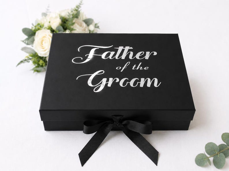 May include: A rectangular black gift box featuring the silver text Father of the Groom printed on the lid. The box is secured with a black ribbon tied in a bow at the front center. The design is clean and simple with a matte finish on the box surface.