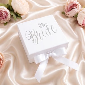 May include: A square white gift box featuring the word Bride in silver script lettering with a small diamond icon above it. The box is closed with a white satin ribbon tied in a bow at the front, surrounded by several soft pink peony flowers.