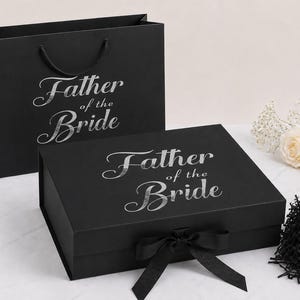 May include: A black gift bag and a matching black rectangular gift box, both featuring the silver text "Father of the Bride" in a script font. The box includes a black ribbon closure, and there is a pile of black shredded paper nearby.