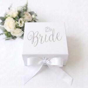 May include: A square white gift box featuring the word Bride in silver script lettering with a small diamond icon above it. The box is closed with a white satin ribbon tied in a bow at the front, accompanied by a small bouquet of white roses.