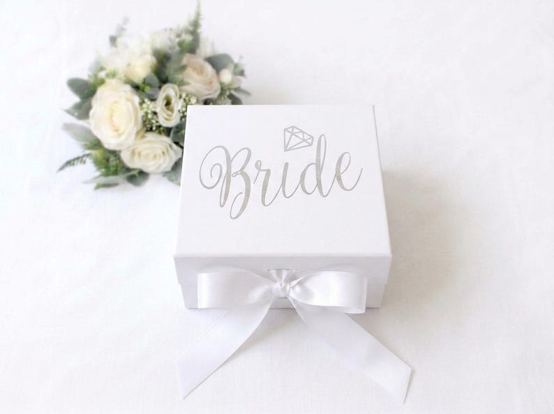 May include: A square white gift box featuring the word Bride in silver script lettering with a small diamond icon above it. The box is closed with a white satin ribbon tied in a bow at the front, accompanied by a small bouquet of white roses.