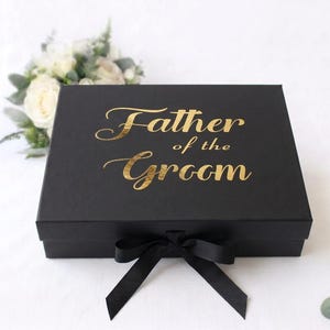 May include: A rectangular black gift box featuring the gold foil text Father of the Groom printed on the lid. The box is secured with a black satin ribbon tied in a bow at the front center, providing a sleek and elegant presentation for gifts.