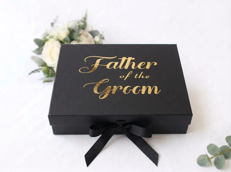May include: A rectangular black gift box featuring the gold foil text Father of the Groom printed on the lid. The box is secured with a black satin ribbon tied in a bow at the front center, providing a sleek and elegant presentation for gifts.