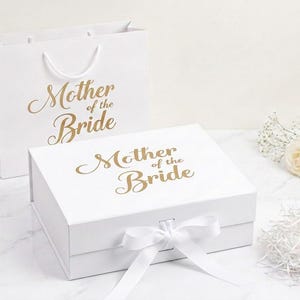 May include: A white gift bag and a rectangular white gift box, both featuring the gold text Mother of the Bride. The box has a white ribbon tied in a bow at the front centre. Two white roses and a small pile of white paper shred are nearby.