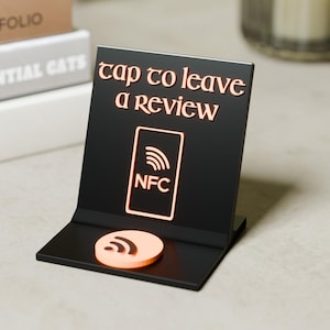 Può includere: Un cartello nero a forma di L con testo color rame che recita Tap to leave a review. Il cartello presenta un'icona rettangolare color rame con un simbolo NFC e un elemento base circolare color rame con un'icona di segnale wireless.