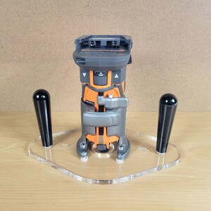 Pu&ograve; includere: Una fresatrice cordless nera e arancione con una base in acrilico trasparente e due manici neri. La fresatrice &egrave; montata sulla base e i manici sono fissati ai lati della base.