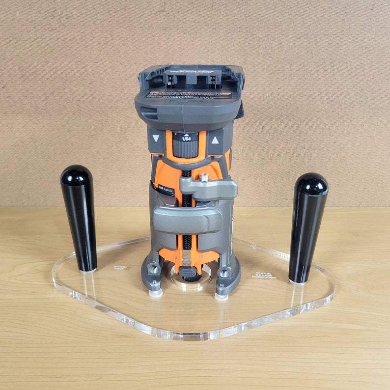 Pu&ograve; includere: Una fresatrice cordless nera e arancione con una base in acrilico trasparente e due manici neri. La fresatrice &egrave; montata sulla base e i manici sono fissati ai lati della base.