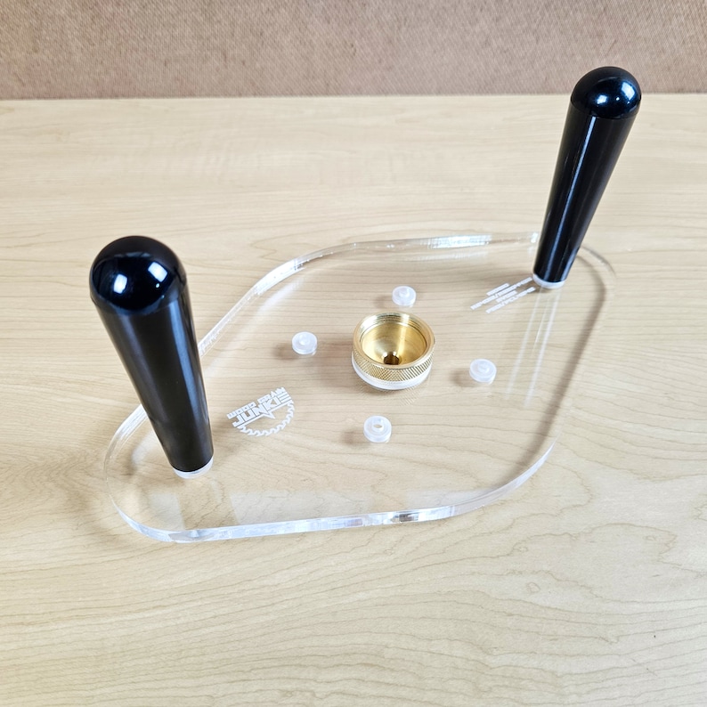 Pu&ograve; includere: Un modello di router in plastica trasparente con due manici neri e un pezzo centrale in ottone. Il modello ha le parole "Wood Grain Junkie" stampate su di esso.