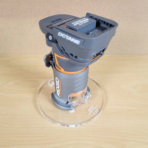 Puede incluir: Una fresadora inal&aacute;mbrica Ridgid gris y naranja con una base de pl&aacute;stico transparente. La fresadora est&aacute; sobre una base de acr&iacute;lico transparente. La fresadora est&aacute; optimizada para bater&iacute;as Octane.