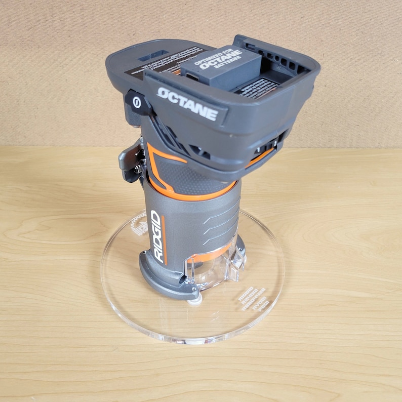 Puede incluir: Una fresadora inal&aacute;mbrica Ridgid gris y naranja con una base de pl&aacute;stico transparente. La fresadora est&aacute; sobre una base de acr&iacute;lico transparente. La fresadora est&aacute; optimizada para bater&iacute;as Octane.