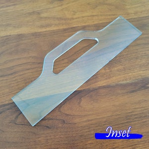 May include: A clear plastic template with a handle cutout. The template is designed for creating an inset, as indicated by the word "Inset" printed in blue on the wooden surface.