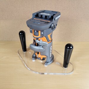 Pu&ograve; includere: Una fresatrice cordless nera e arancione con una base in acrilico trasparente e due manici neri. La fresatrice &egrave; etichettata "RIDGID OCTANE".