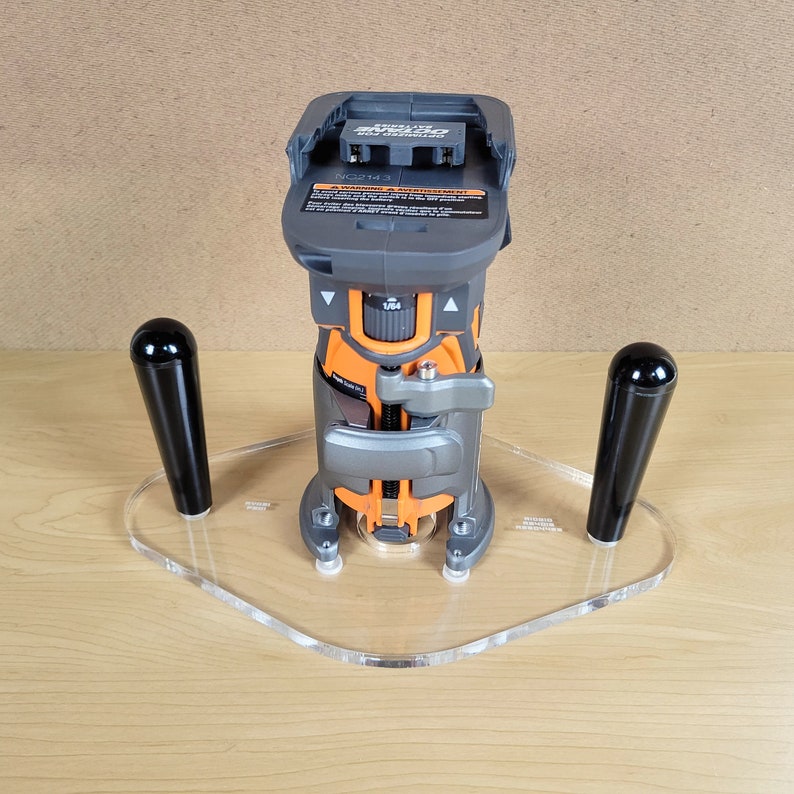 Pu&ograve; includere: Una fresatrice cordless grigia e arancione con una batteria collegata. La fresatrice &egrave; montata su una base in acrilico trasparente con due maniglie nere. La base ha la scritta "ROUTER BASE" stampata.