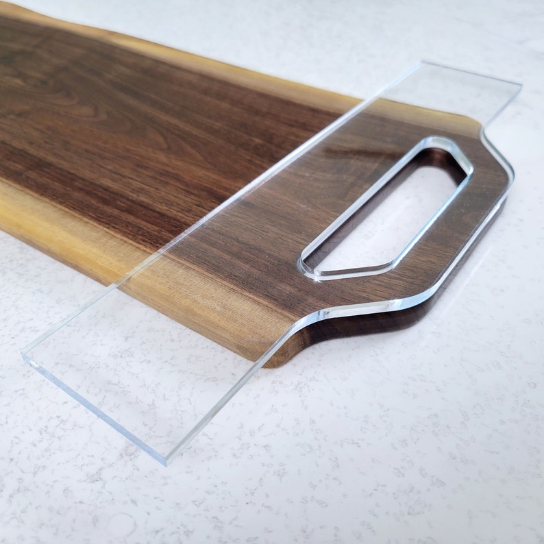 May include: A wooden serving board with a clear acrylic handle. The board is made of dark wood with a natural edge. The handle is shaped like a loop and has a smooth, polished finish.