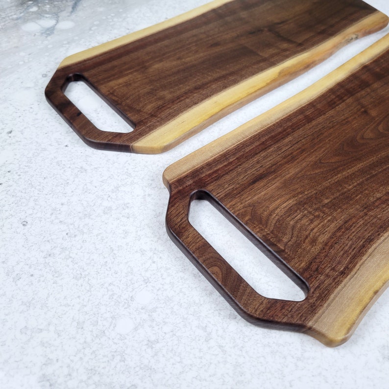 May include: Two wooden serving boards with handles. The boards are made of walnut wood and have a natural edge. The boards are both rectangular in shape and have a light brown color.