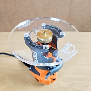 Puede incluir: Una plantilla de router de pl&aacute;stico transparente con un inserto de lat&oacute;n y un router negro y naranja adjunto. La plantilla tiene el texto "Woodcraft" y "Made in China" impreso en ella.