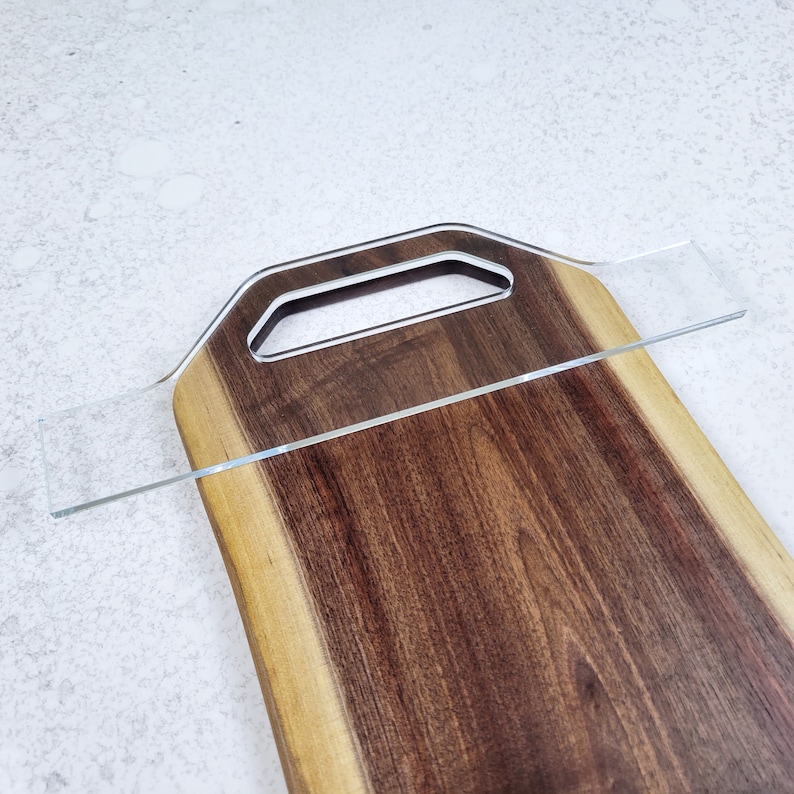 May include: A large wooden cutting board with a natural edge and a handle. The board is made of dark wood with a lighter wood border. A clear acrylic piece is placed on top of the board.