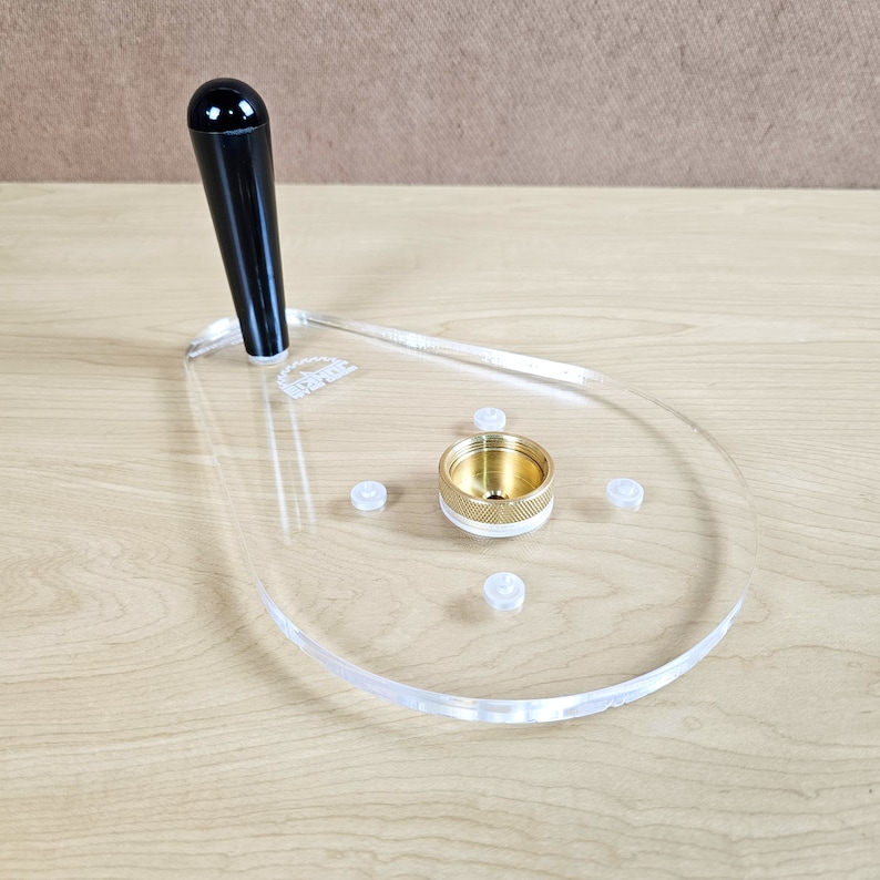 Puede incluir: Un jig de router de acr&iacute;lico transparente con un mango negro y un inserto de lat&oacute;n. El jig tiene cuatro peque&ntilde;os pies de pl&aacute;stico blanco.