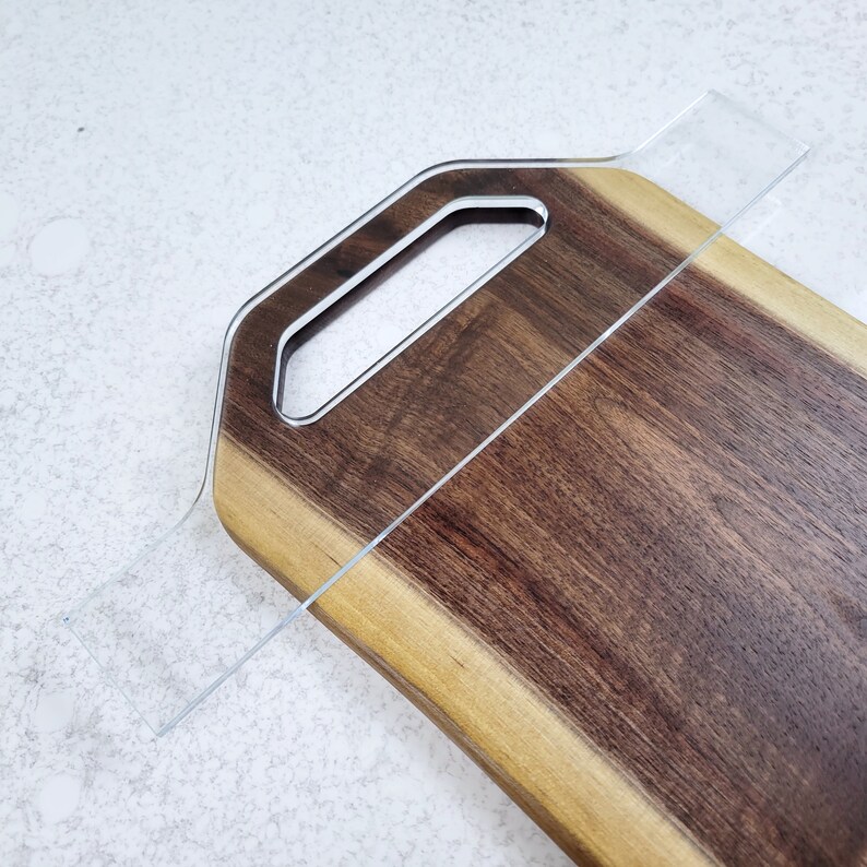May include: A wooden cutting board with a natural edge and a handle. The board is made of walnut wood and has a light brown color. A clear acrylic ruler is placed on top of the board.