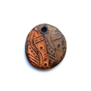 May include: A small, round, ceramic pendant with a hole at the top. The pendant is divided into two sections, one side is a reddish-brown color with a dotted and curved design, and the other side is a darker brown with a linear pattern.