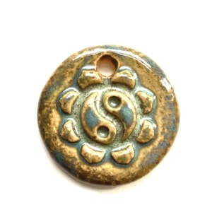 May include: A round ceramic pendant with a blue and white yin yang symbol surrounded by a floral design. The pendant is glazed in shades of brown and green.