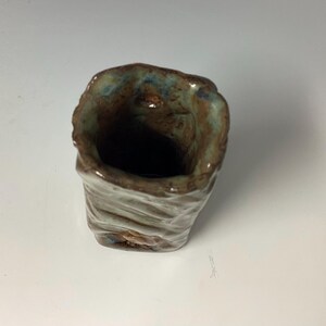 May include: A small, handmade ceramic cup with a textured, brown, green, and blue glaze. The cup has a wide opening and a rounded bottom.