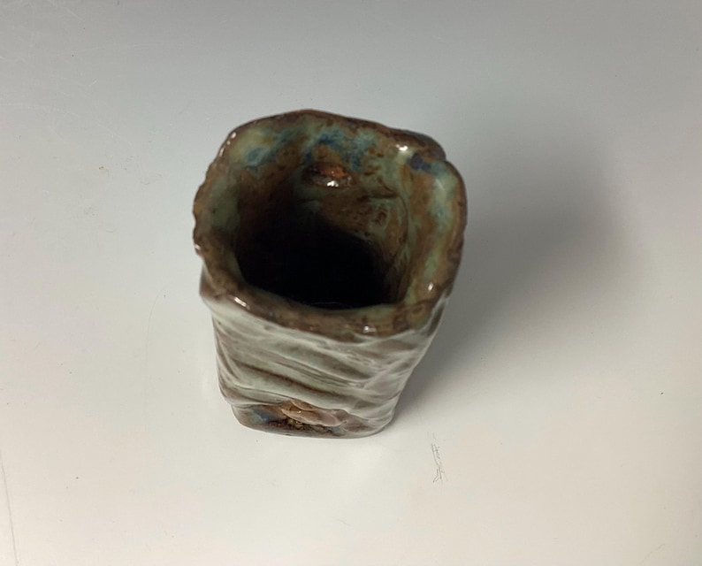 May include: A small, handmade ceramic cup with a textured, brown, green, and blue glaze. The cup has a wide opening and a rounded bottom.